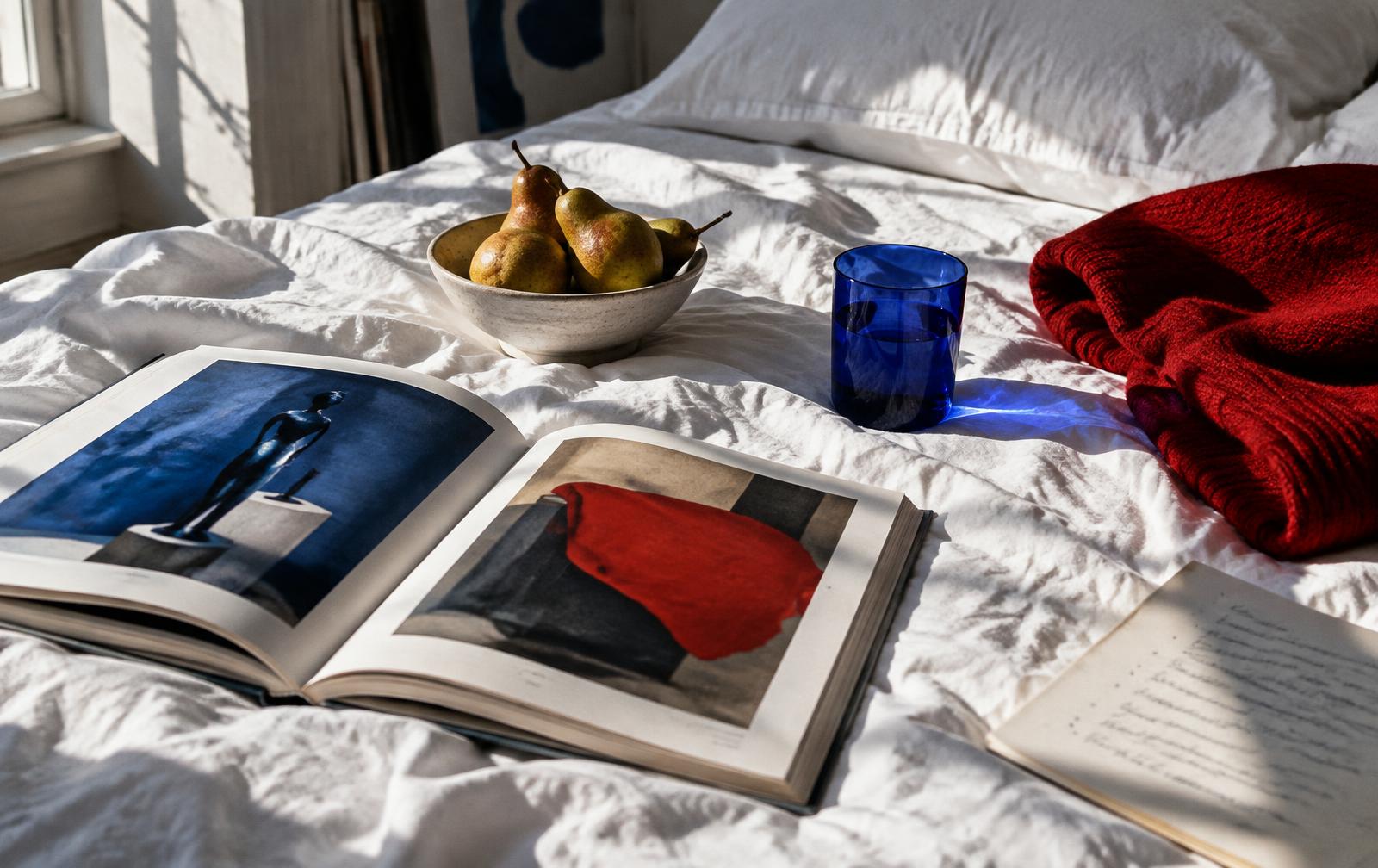 Art books, pears, a blue glass and a red knit on a sunlit bed